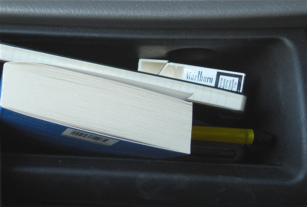 An open car glovebox holding a paperback book, a notebook, a cigarette pack, and a pen.